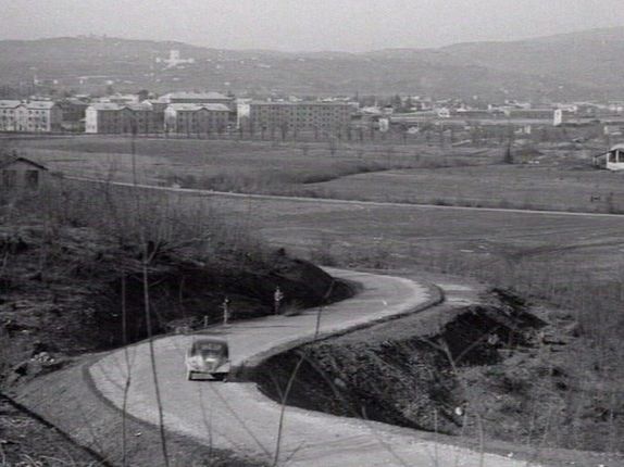 . The road through Rafut shortly after construction. In the background, the railway workers’ housing blocks and the Oslavia ossuary. &copy;still from the documentary &quot