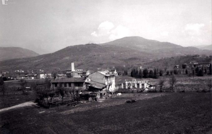 Fotografija hiše, preden so jo porušili z ozadjem. Vidne tudi sosednje hiše. Domnevno med 1953 in 1955. Na kmetiji zadaj so imeli prašiče..  